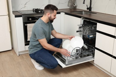 Photo of Smiling man loading dishwasher with plates in kitchen