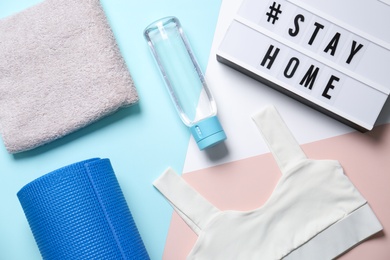 Photo of Stylish sportswear, yoga mat and light box with hashtag Stay at Home on color background, flat lay. Self isolation during COVID‑19 pandemic