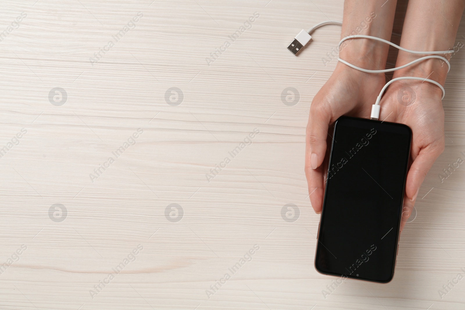 Photo of Internet addiction. Top view of woman holding phone at wooden table, hand tied to device with charging cable