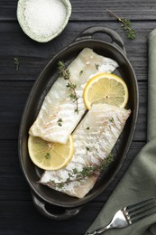 Photo of Fresh raw cod fillets with thyme and lemon in baking dish on black wooden table, flat lay