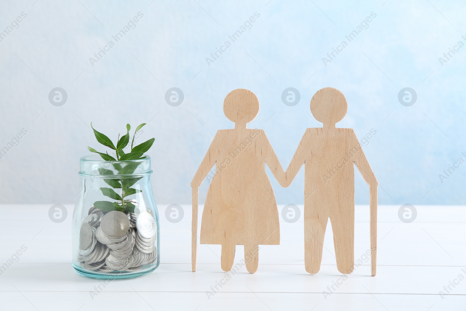 Photo of Pension savings. Figure of senior couple, jar of coins and green twig on white wooden table