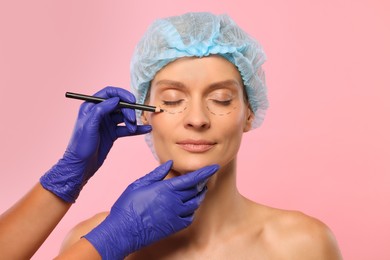 Doctor with pencil preparing patient for cosmetic surgery operation on pink background