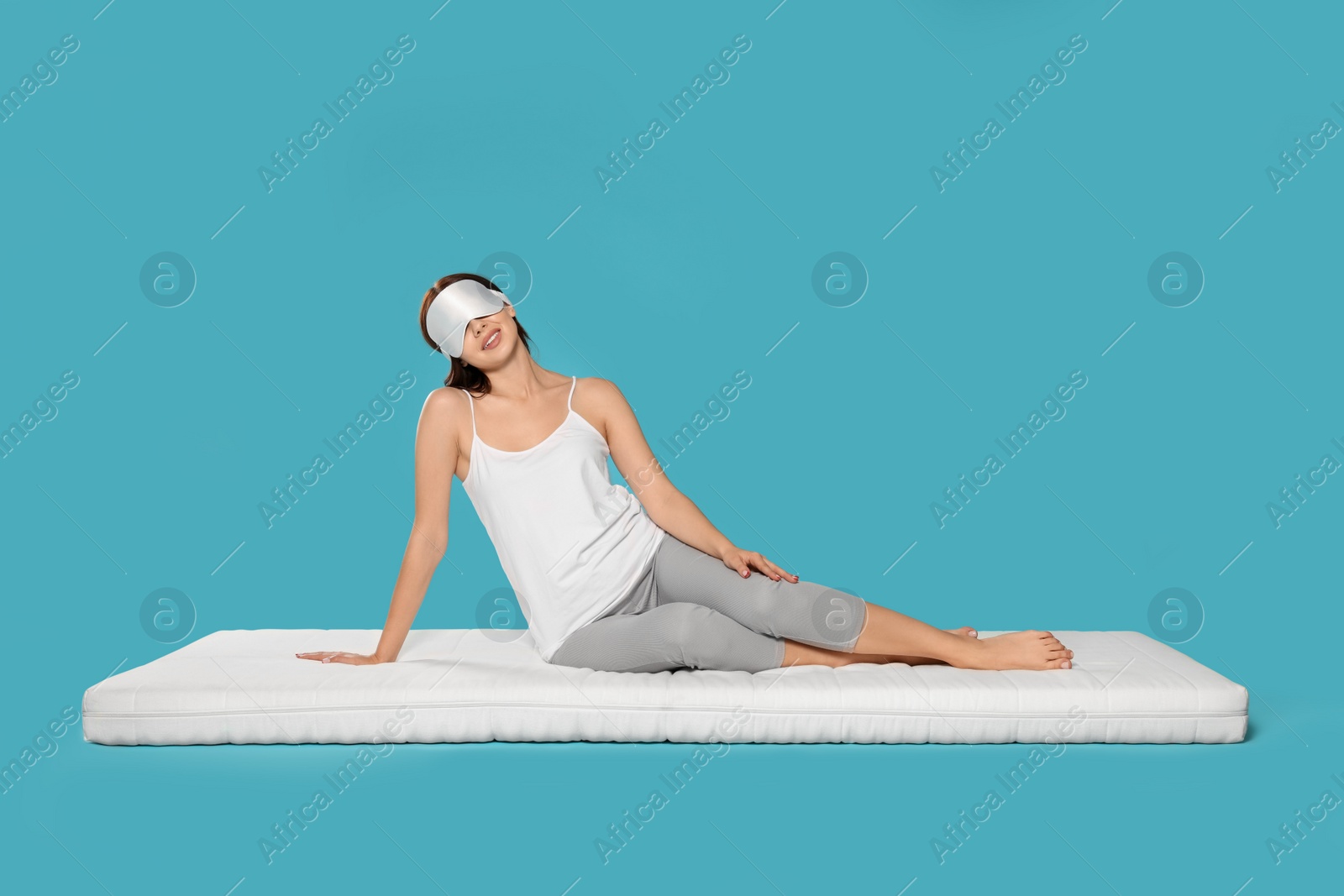 Photo of Woman in sleep mask sitting on soft mattress against light blue background