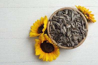 Raw sunflower seeds in bowl and flowers on white wooden table, top view