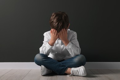 Photo of Upset boy sitting on floor near black wall. School bullying