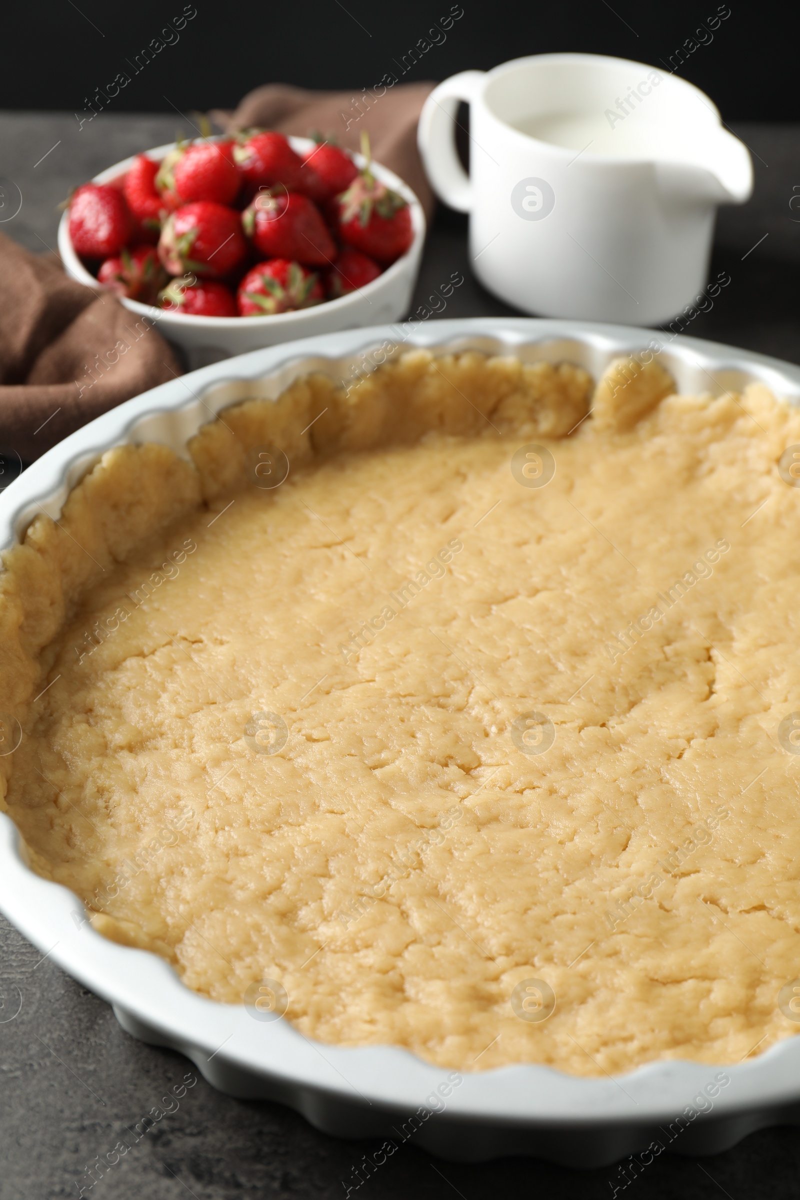 Photo of Making shortcrust pastry. Raw dough in baking dish, strawberries and milk on grey table, closeup