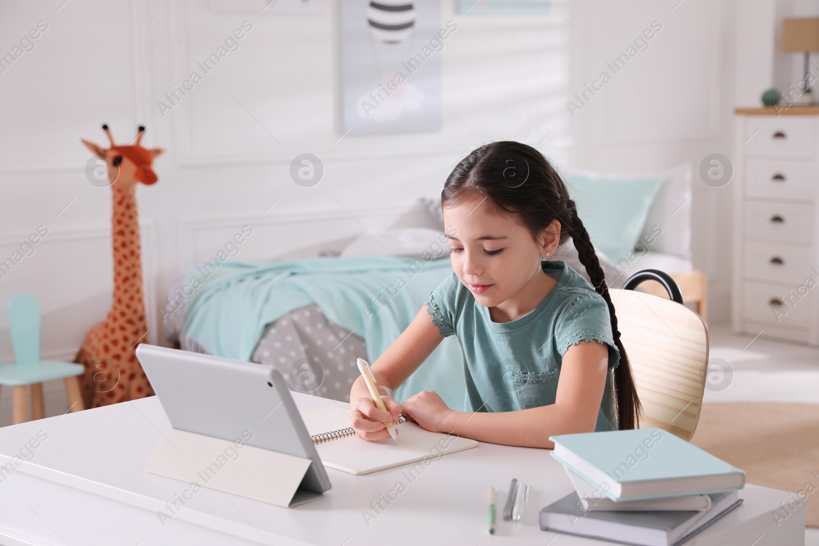 Photo of Little girl doing homework with tablet at table in bedroom