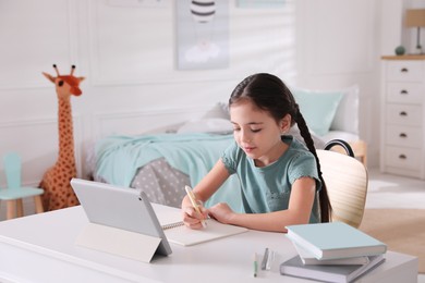 Photo of Little girl doing homework with tablet at table in bedroom