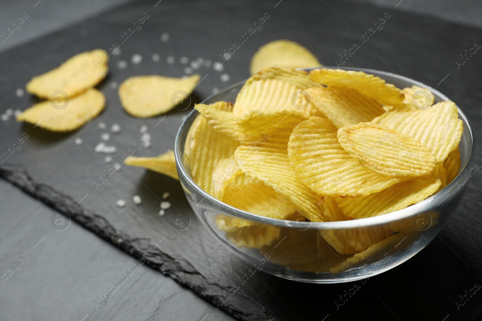 Photo of Slate board with bowl of potato chips on grey table, closeup. Space for text