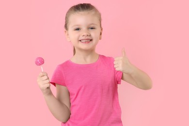 Photo of Happy little girl with lollipop showing thumbs up on pink background