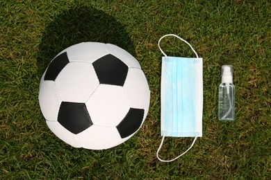 Photo of Football ball with hand sanitizer and protective mask on green grass, flat lay