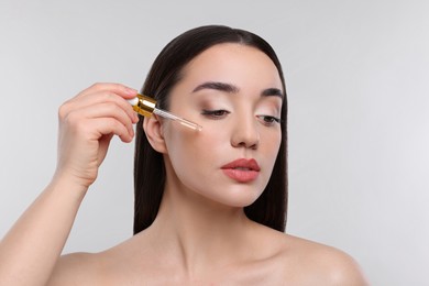 Young woman applying essential oil onto face on light grey background