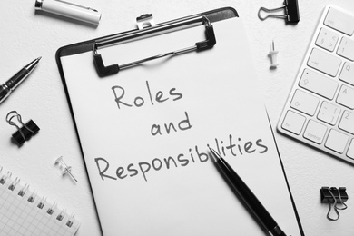 Photo of Clipboard with sheet of paper saying Roles and Responsibilities among office supplies on white table, flat lay