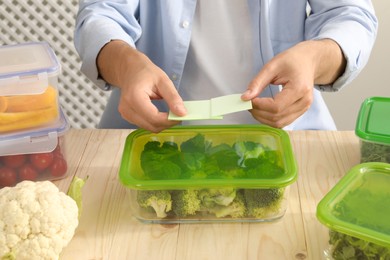 Photo of Man sticking paper note onto container with fresh broccoli at wooden table, closeup. Food storage