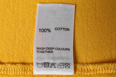 Photo of Clothing label with care symbols and material content on yellow shirt, top view