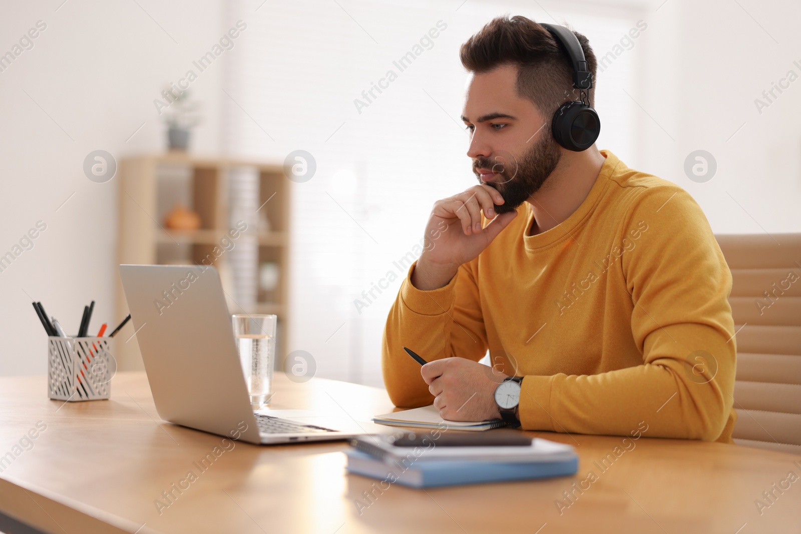 Photo of Young man in headphones watching webinar at table in room