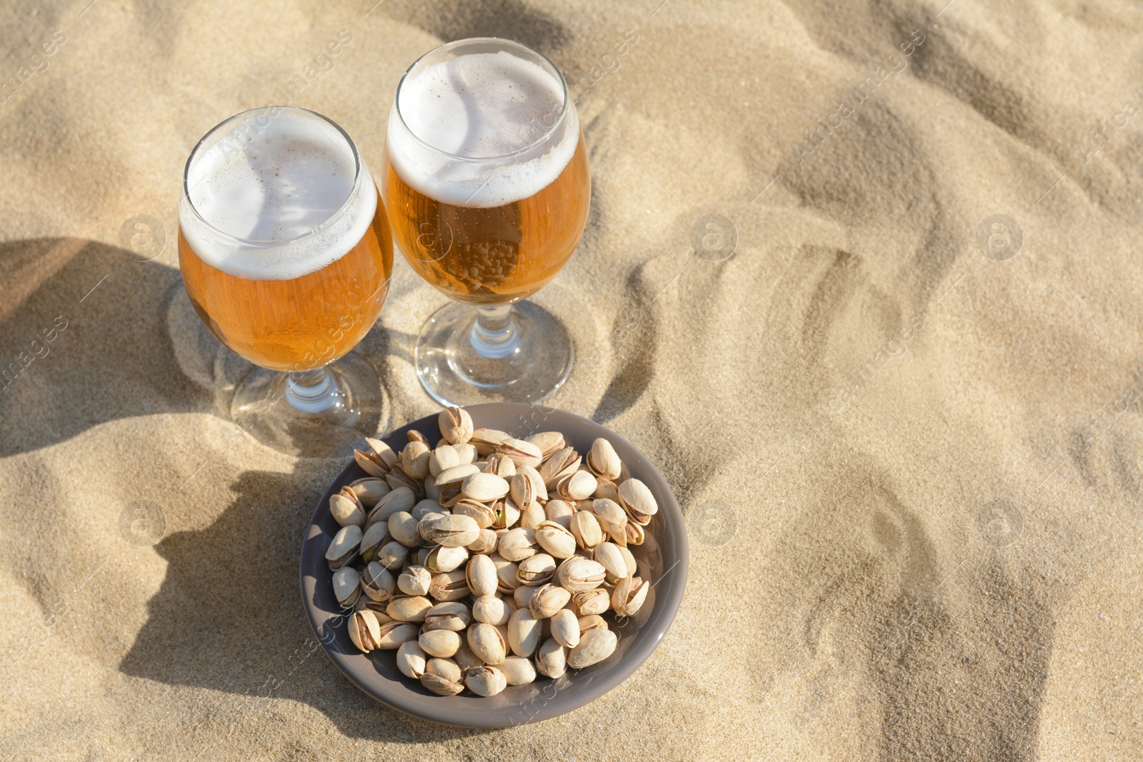 Photo of Glasses of cold beer and pistachios on sandy beach, above view. Space for text