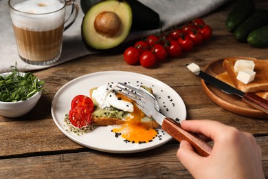 Photo of Woman eating delicious toast with poached egg and avocado at wooden table, closeup