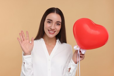 Young woman holding red heart shaped balloon and waving hello on beige background