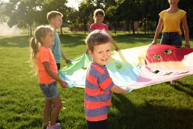 Photo of Group of children and teacher playing with rainbow playground parachute on green grass. Summer camp activity