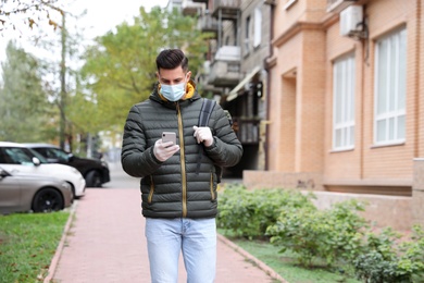 Man in medical face mask and gloves with smartphone walking outdoors. Personal protection during COVID-19 pandemic