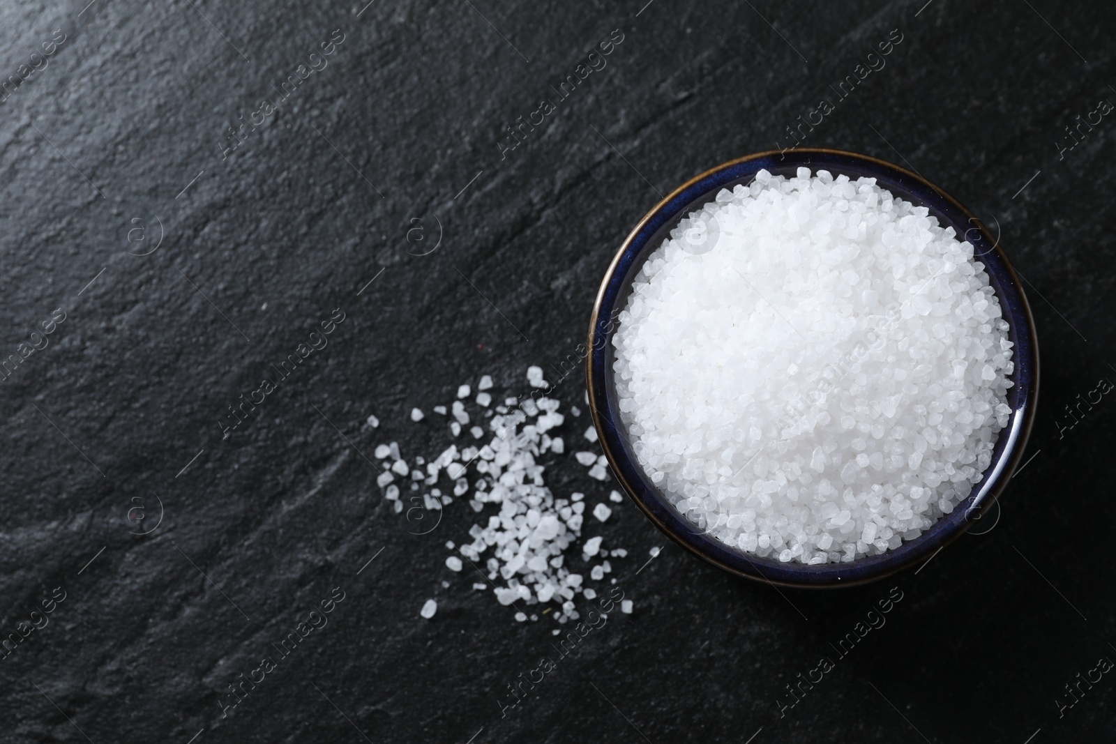 Photo of Organic white salt in bowl on black table, top view. Space for text