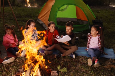 Photo of Little children reading book with flashlight outdoors. Summer camp