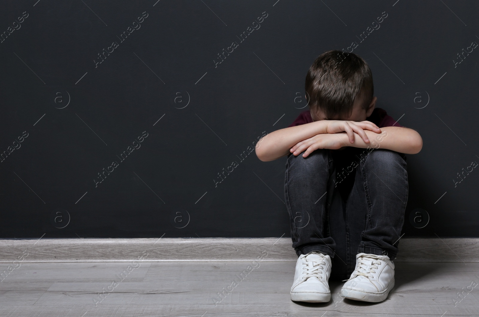 Photo of Sad little boy on floor near black wall. Space for text