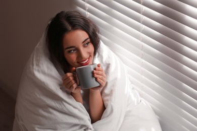 Photo of Beautiful young woman wrapped with soft blanket enjoying hot drink near window at home
