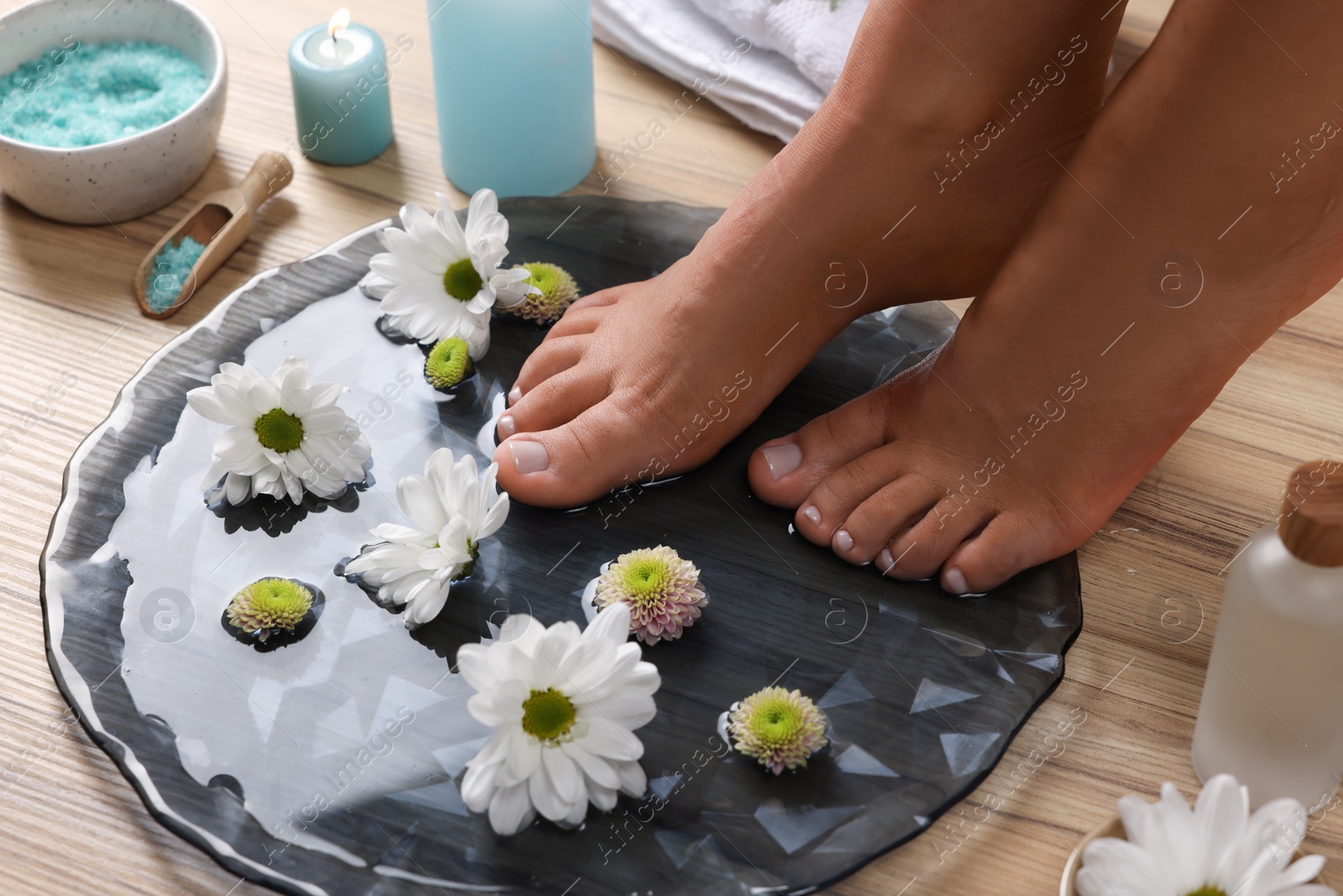 Photo of Woman soaking her feet in plate with water and flowers on wooden floor, closeup. Pedicure procedure