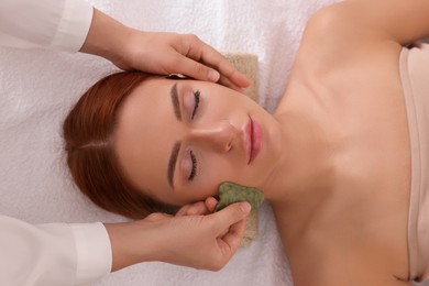 Young woman receiving facial massage with jade gua sha tool in beauty salon, above view