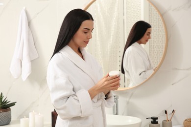 Beautiful young woman with jar of cream near mirror in bathroom