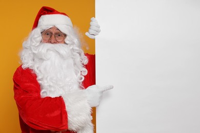 Photo of Santa Claus pointing at blank poster on orange background, space for text