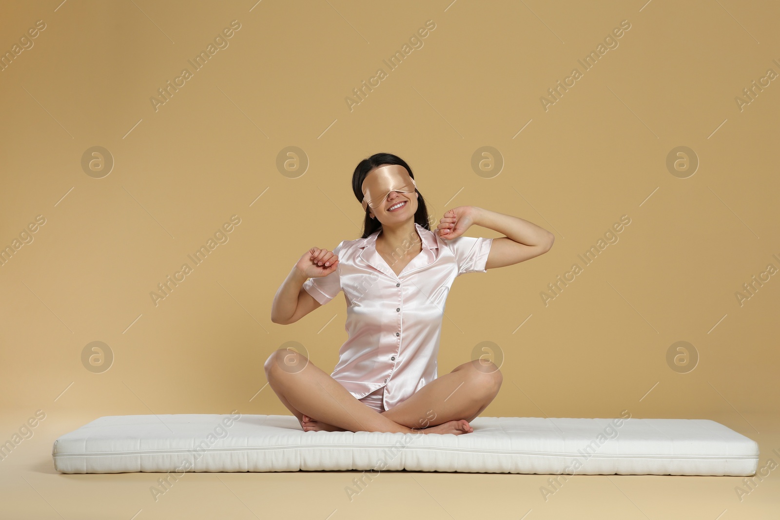 Photo of Woman with sleep mask stretching on soft mattress against beige background