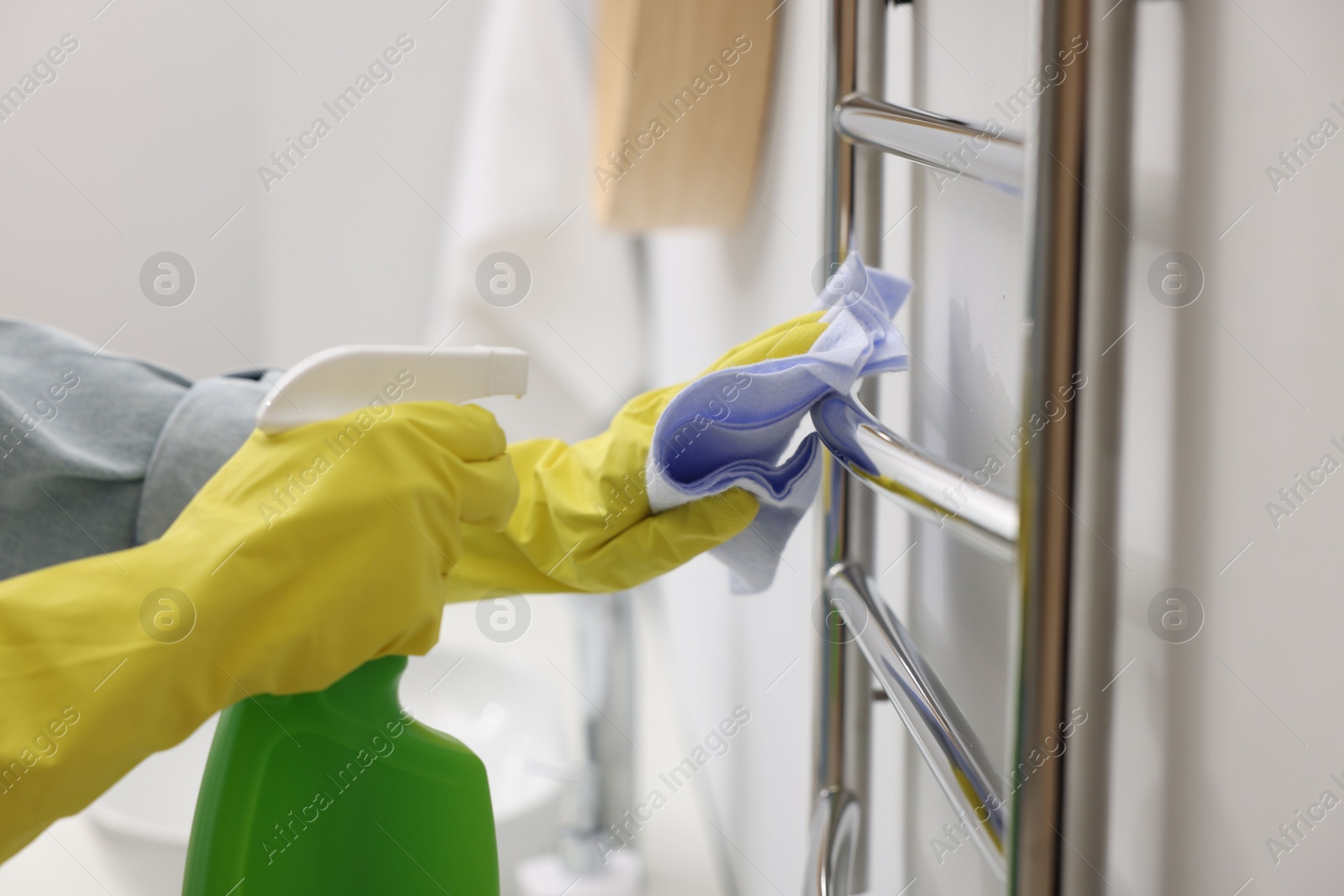 Photo of Woman cleaning heated towel rail with sprayer and rag, closeup