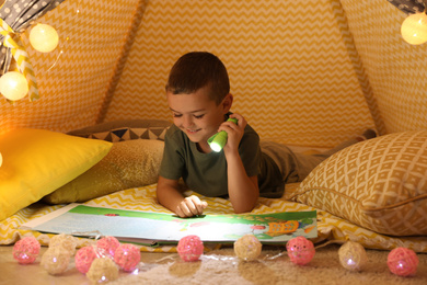 Photo of Little boy with flashlight reading book in play tent
