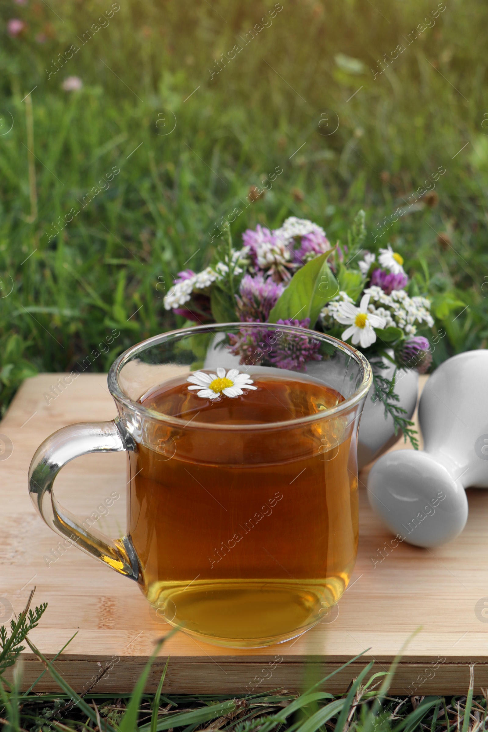 Photo of Cup of aromatic herbal tea, pestle and ceramic mortar with different wildflowers on wooden board in meadow