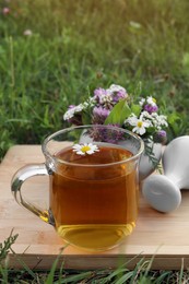 Cup of aromatic herbal tea, pestle and ceramic mortar with different wildflowers on wooden board in meadow
