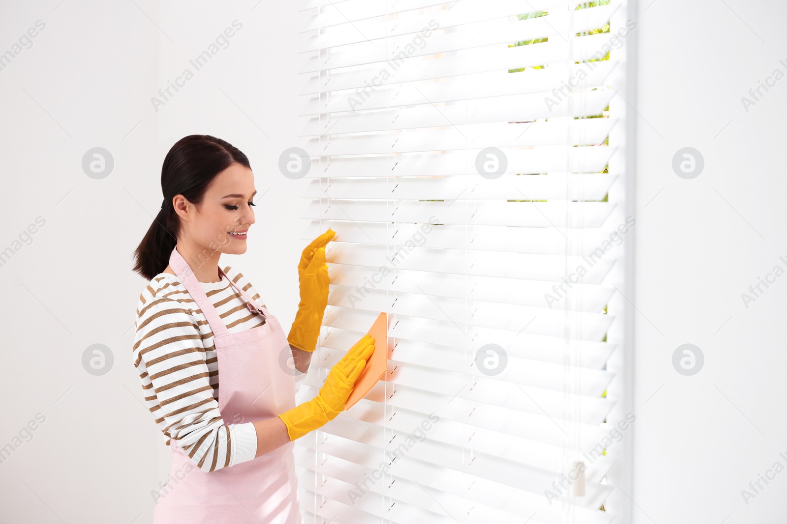 Photo of Young chambermaid wiping dust from blinds in room