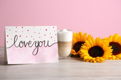Card with phrase Love You, latte macchiato and sunflowers on white wooden table against pink wall