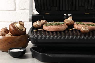 Photo of Electric grill with homemade sausages, rosemary and mushrooms on light table, closeup