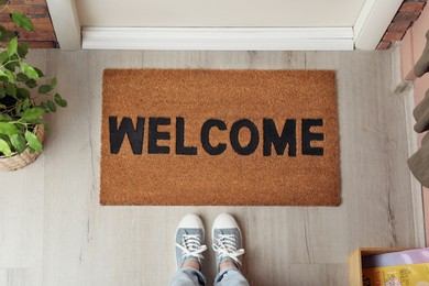 Woman standing near door mat with word Welcome on wooden floor in hall, top view