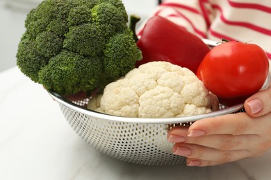 Photo of Woman holding colander with fresh vegetables at white marble table, closeup