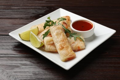 Photo of Tasty fried spring rolls, arugula, lime and sauce on wooden table, closeup