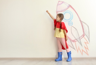 Adorable little child playing astronaut near wall with drawing of spaceship indoors