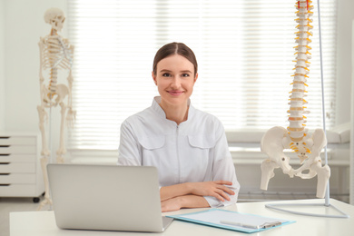 Photo of Female orthopedist with laptop near human spine model in office