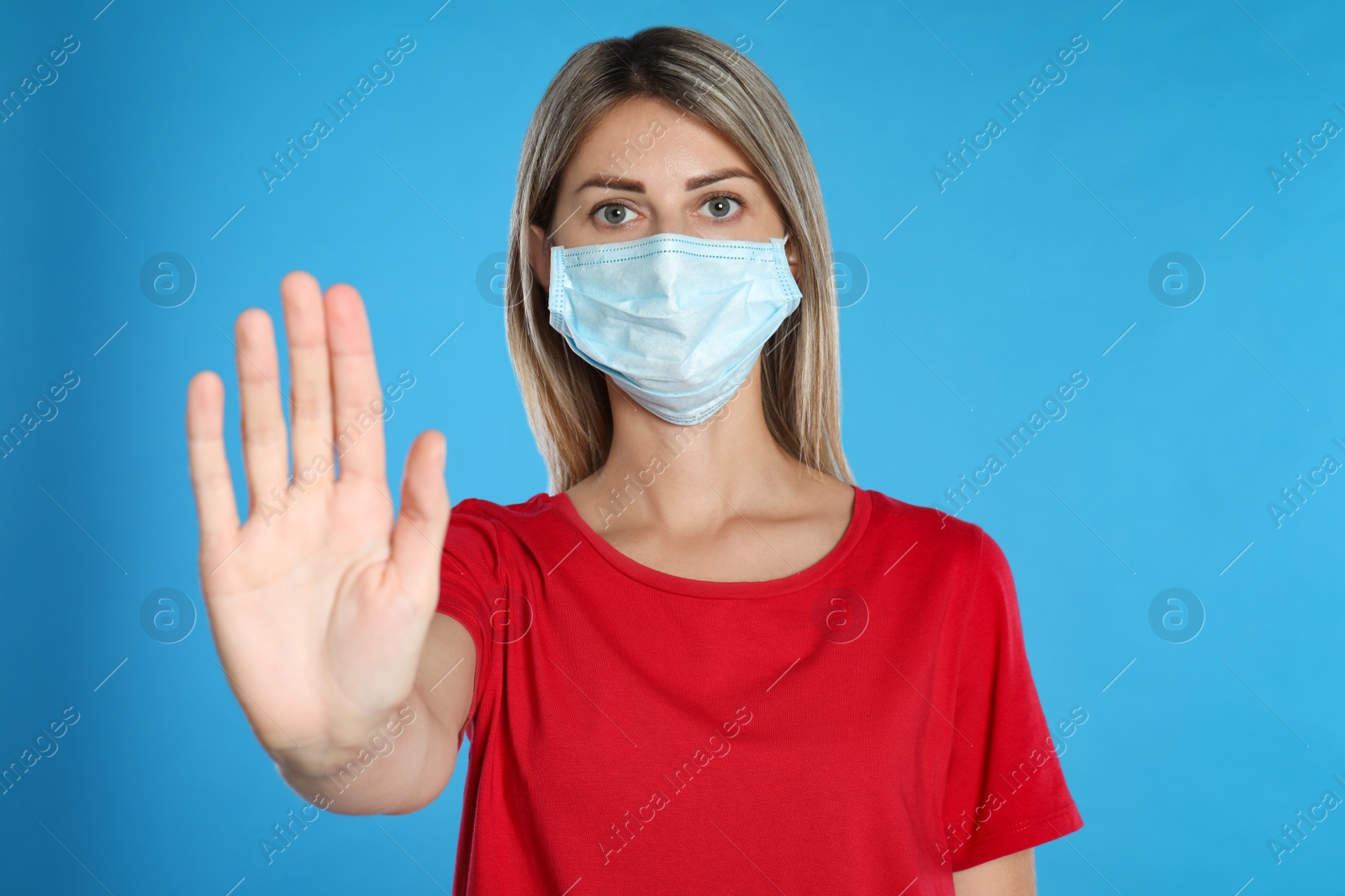 Photo of Woman in protective mask showing stop gesture on light blue background. Prevent spreading of coronavirus