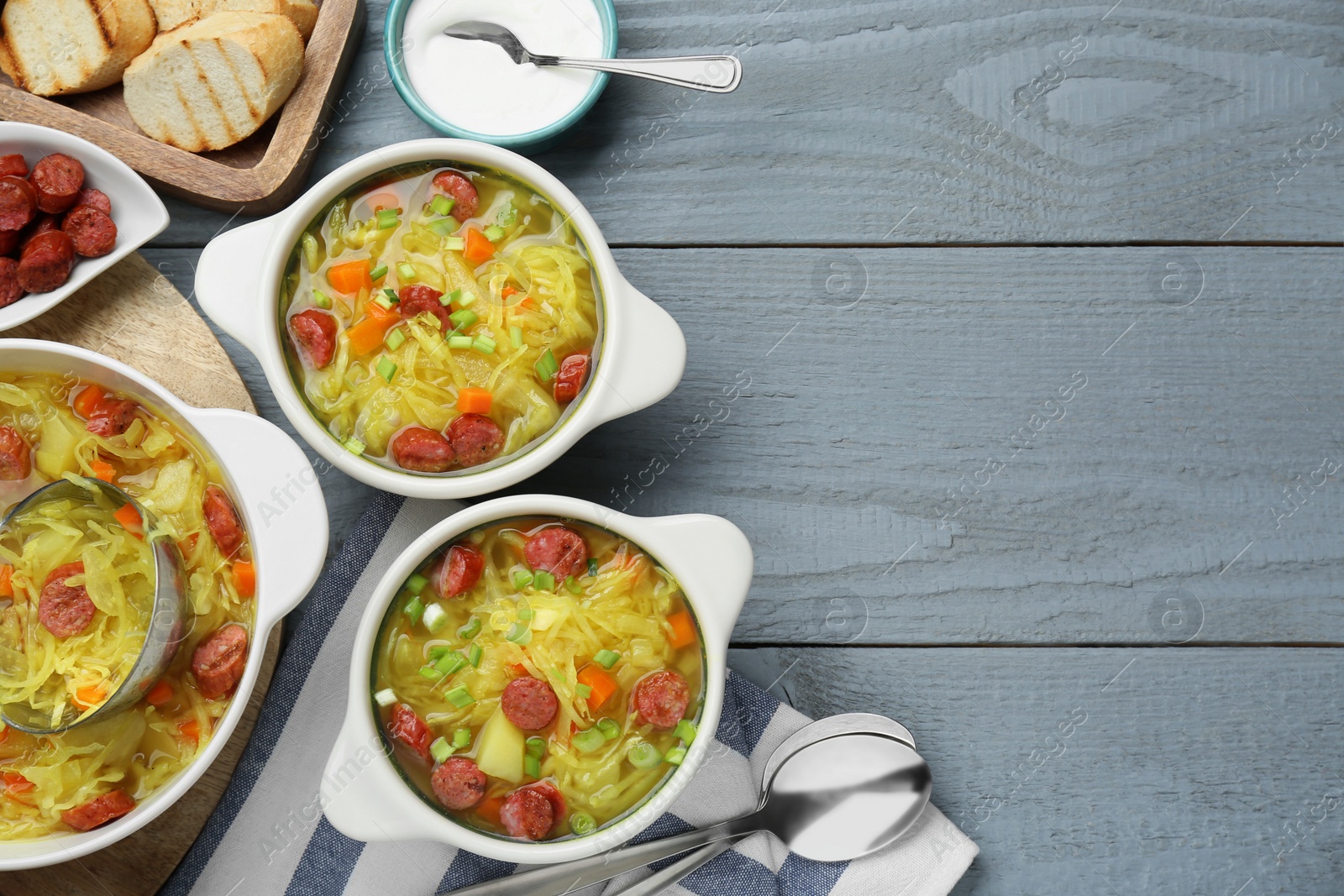 Photo of Delicious sauerkraut soup with smoked sausages and green onion served on grey wooden table, flat lay. Space for text