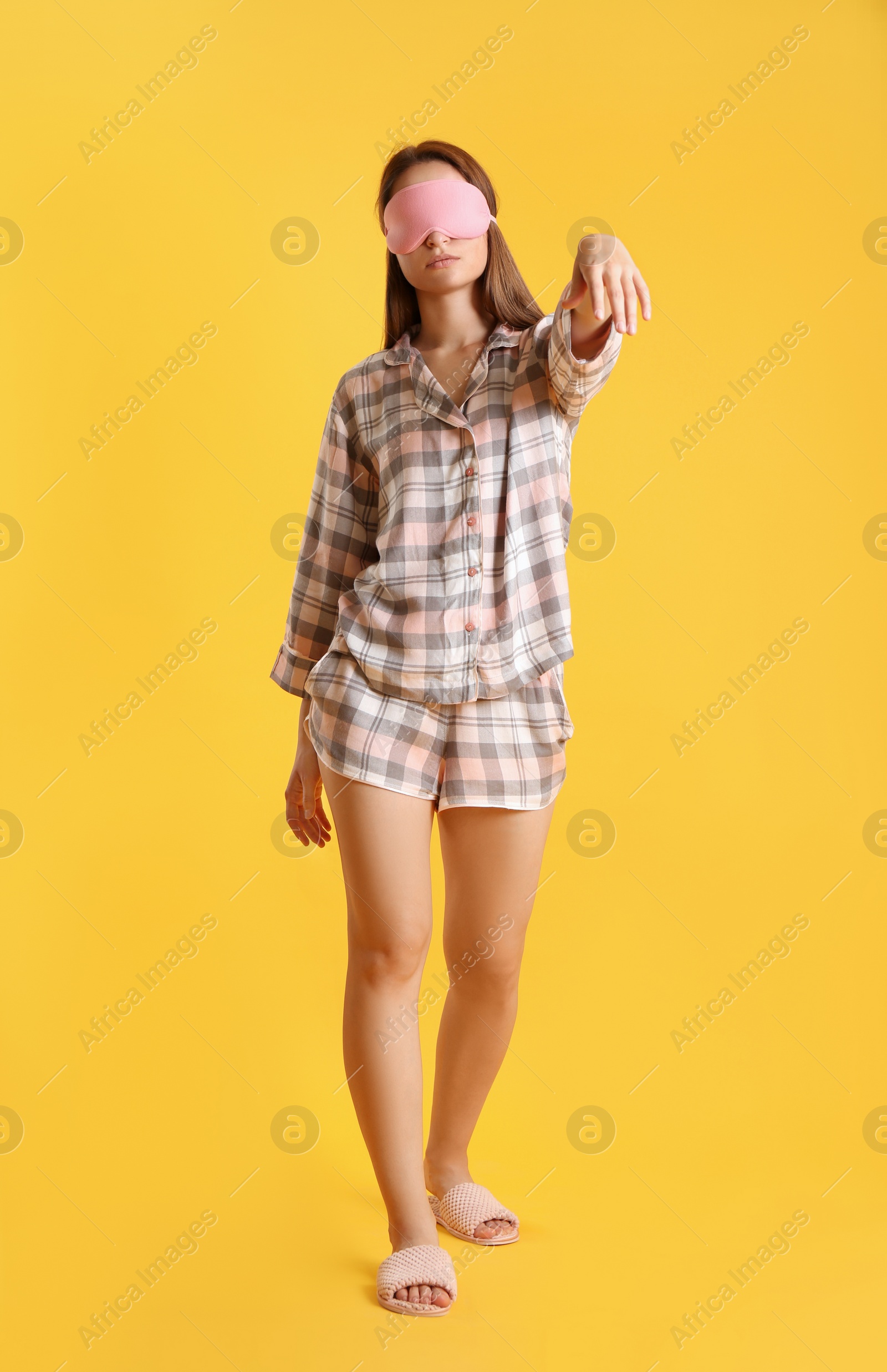 Photo of Young woman wearing pajamas, mask and slippers in sleepwalking state on yellow background