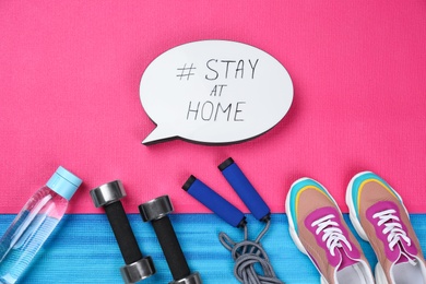 Photo of Sport equipment and speech bubble with hashtag STAY AT HOME on colorful yoga mats, flat lay. Message to promote self-isolation during COVID‑19 pandemic